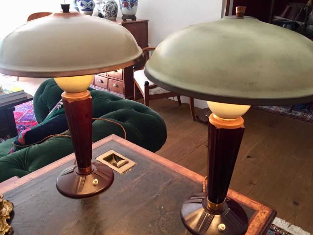 A near pair of Art Deco desk lamps featuring stepped dark brown Bakelite bases, fluted stems, and wide rounded metal shades with internal illuminated glass diffusers.