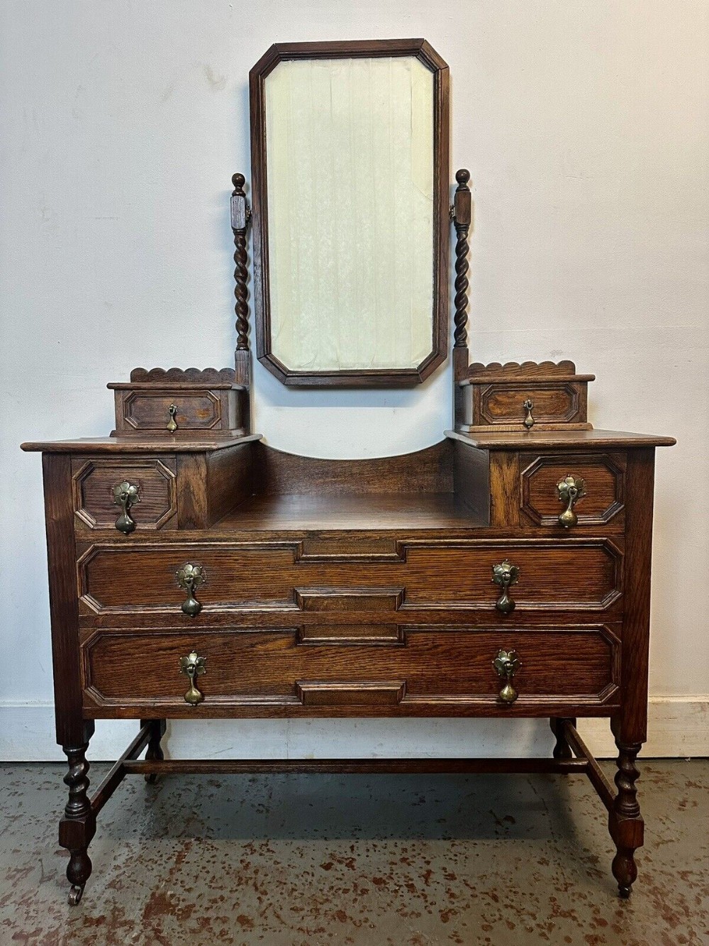 Dark oak dressing table chest featuring geometric panelled drawers, brass drop handles, small jewel drawers, and a central swinging mirror supported by barley twist columns.