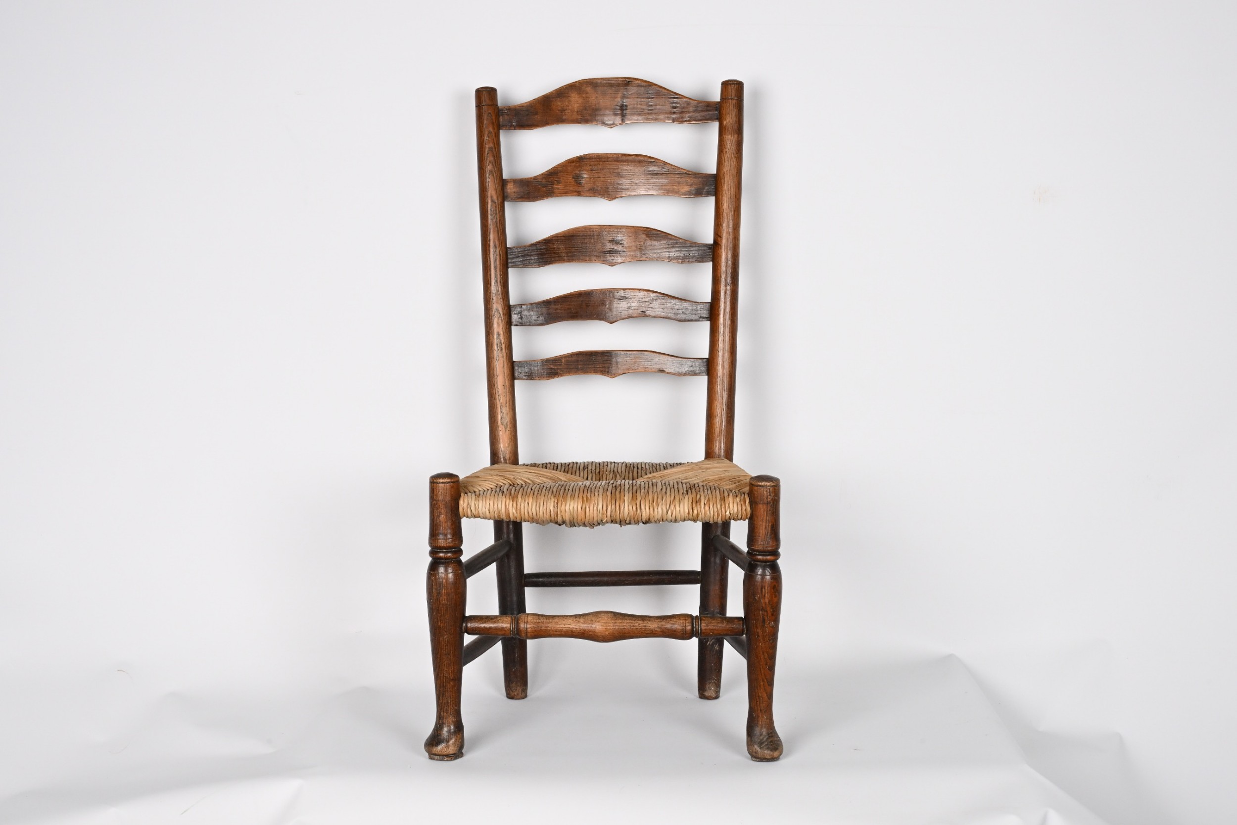 An antique dark elm ladder back chair featuring a woven rush seat and turned front legs with stretchers.