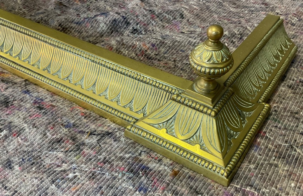 An antique Victorian brass fireplace fender featuring a low rectangular kerb with decorative fluted moulding and detailed finials at each end. View V