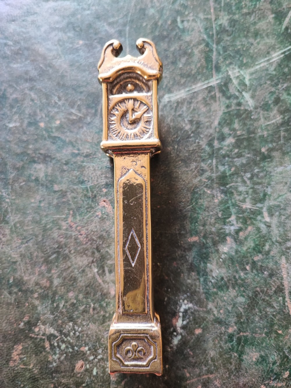 A small cast brass novelty door knocker shaped like a miniature grandfather clock with a detailed dial and pedestal base. View X