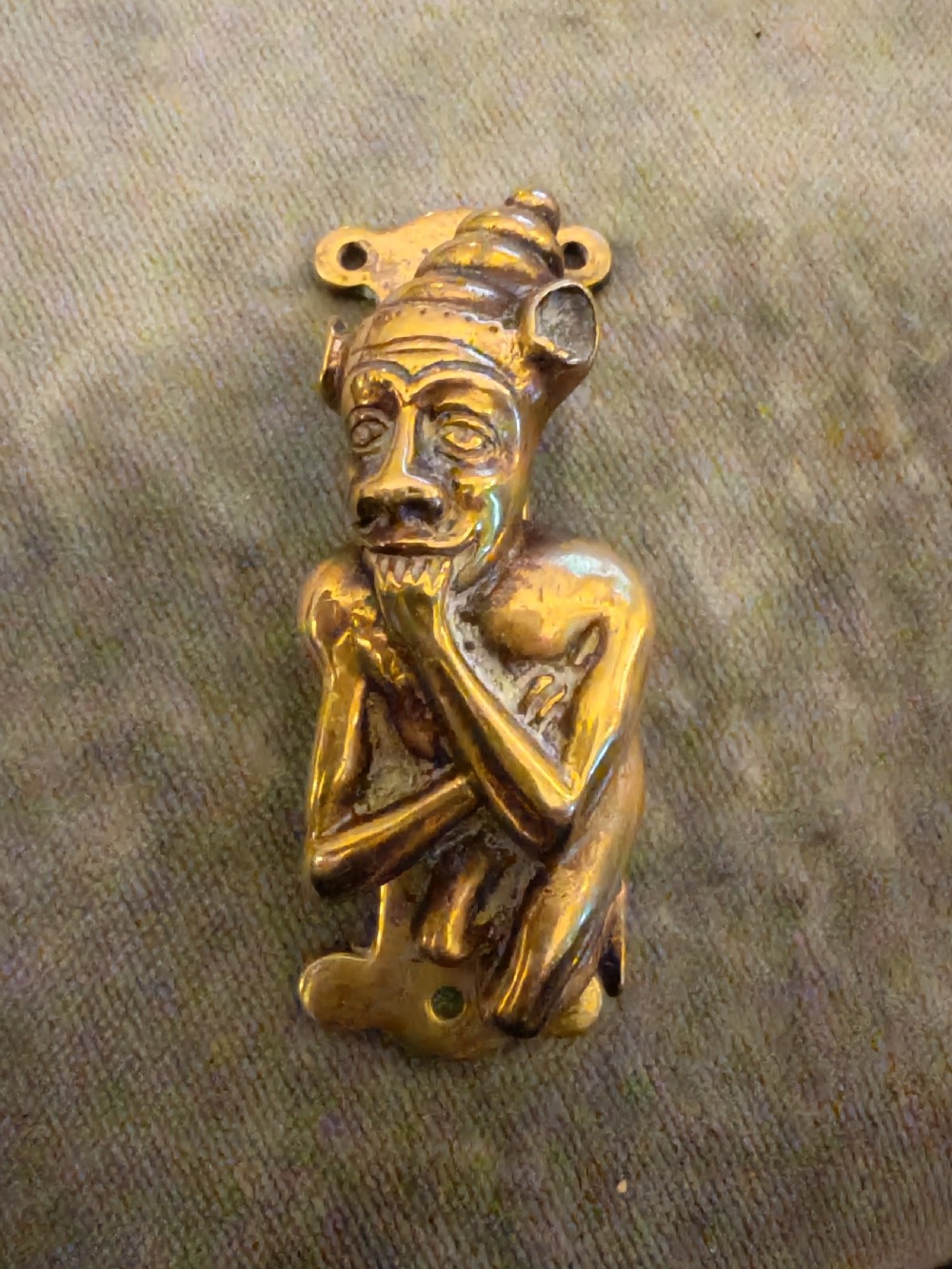 An antique cast brass door knocker depicting a seated imp figure resting its chin on its hand with mounting holes at the top and bottom.