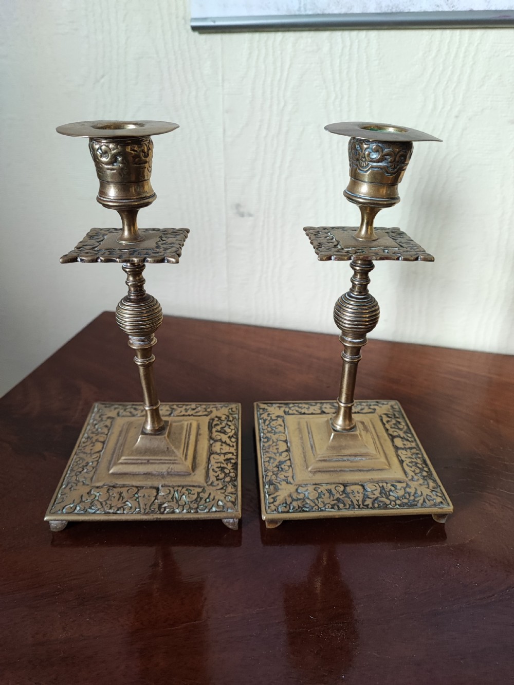 A matching pair of Victorian brass candlesticks featuring square relief bases, ribbed stems, square drip trays, and turned sconces.