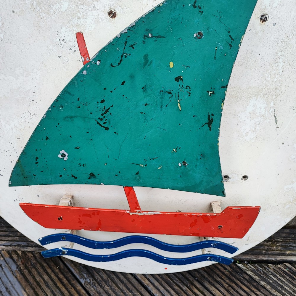 A circular white painted wooden wall sign featuring a raised relief motif of a sailboat with a green sail, red hull, and blue wave lines. View II