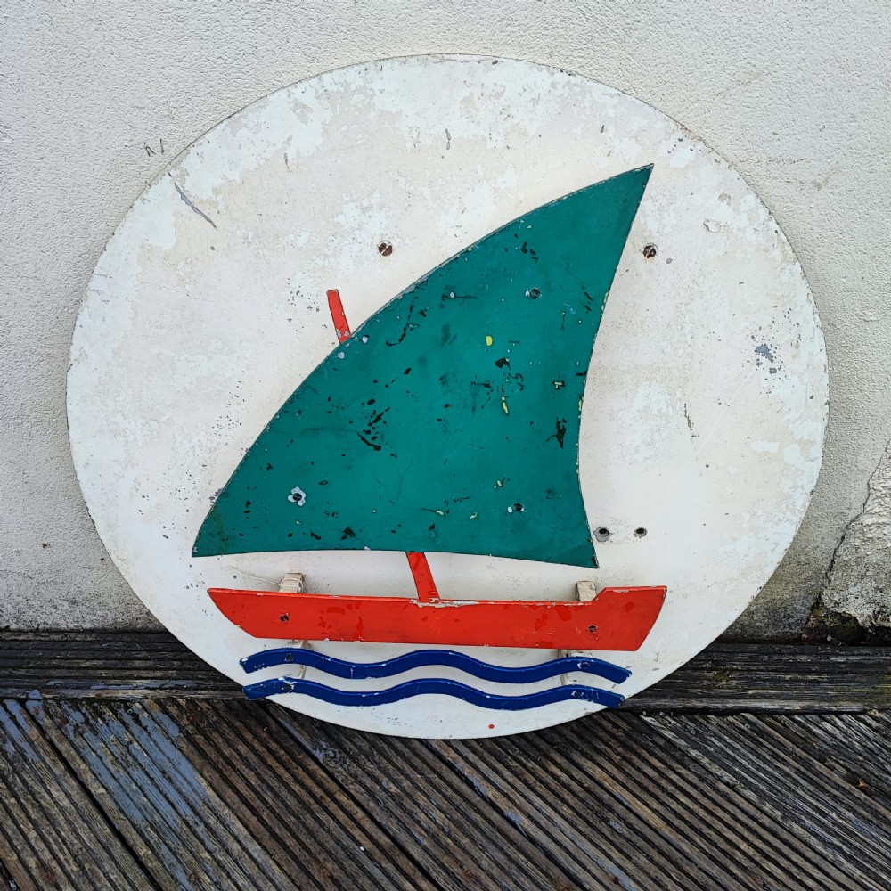 A circular white painted wooden wall sign featuring a raised relief motif of a sailboat with a green sail, red hull, and blue wave lines. View IV
