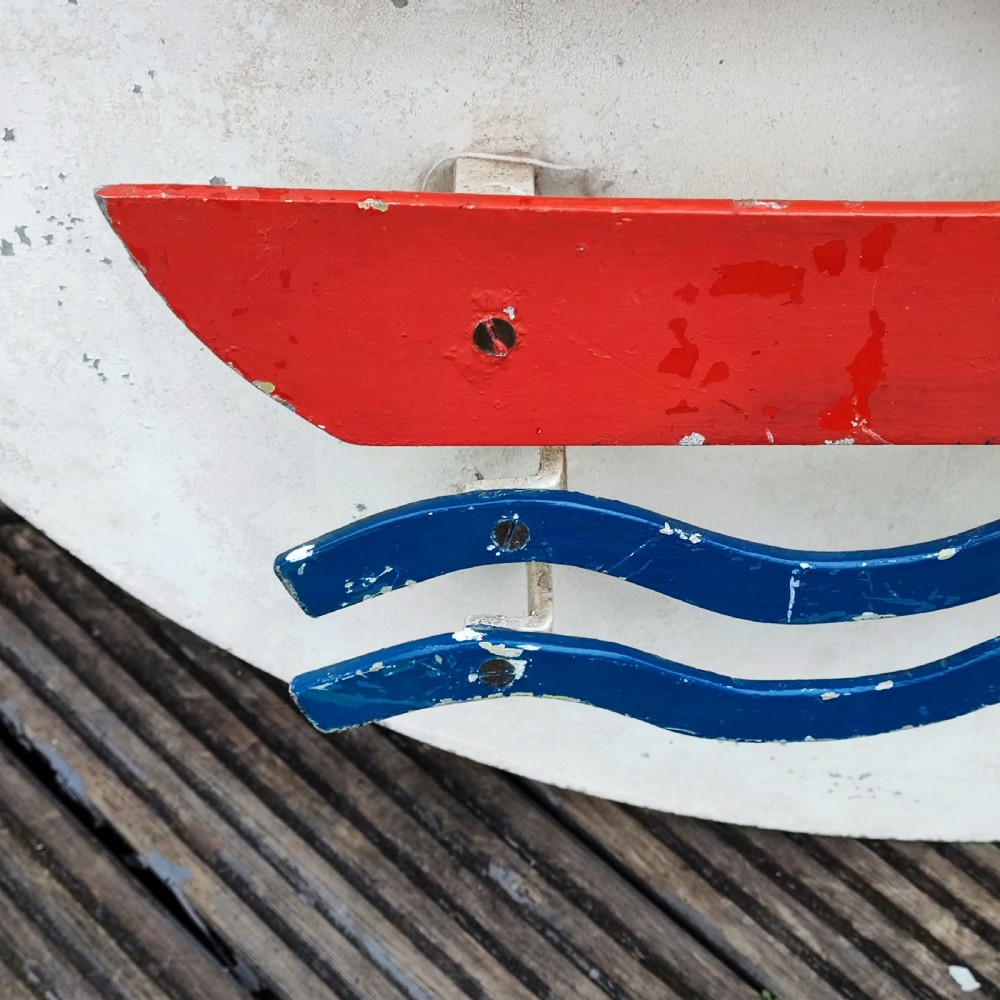 A circular white painted wooden wall sign featuring a raised relief motif of a sailboat with a green sail, red hull, and blue wave lines. View V