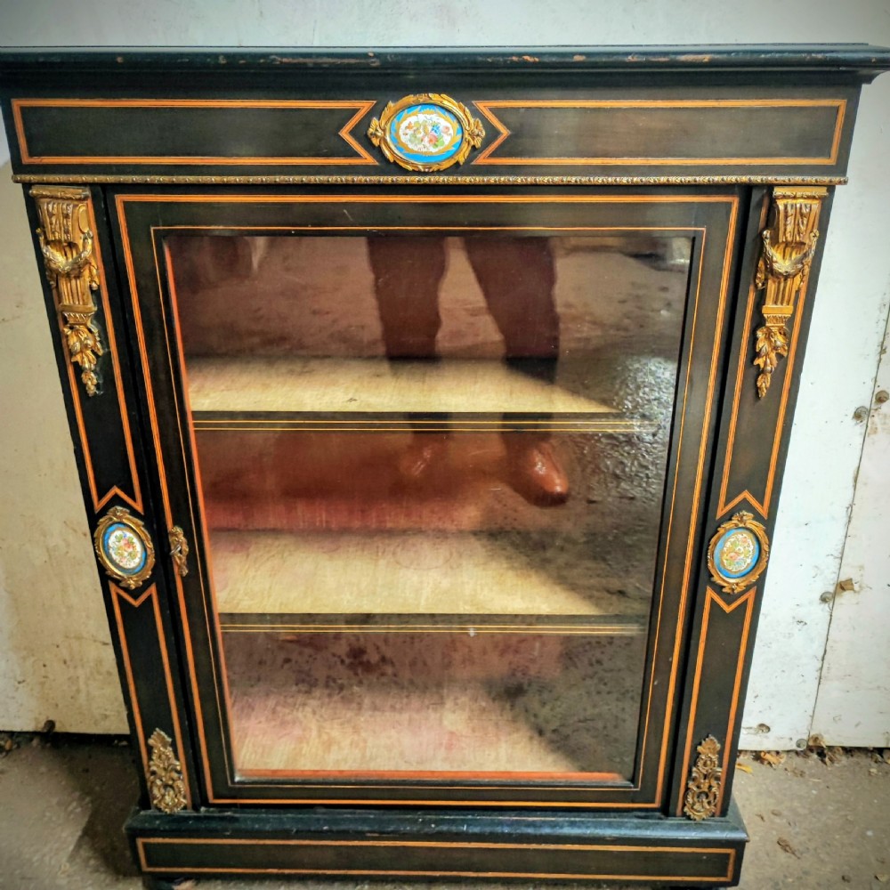 A black ebonised Victorian display cabinet featuring a central glazed door, gilt metal mounts, stringing inlay, and three porcelain plaques.