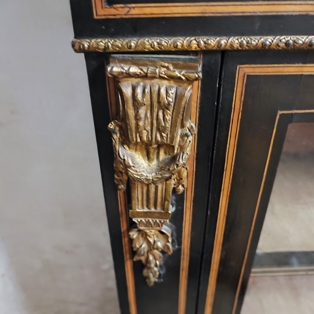A black ebonised Victorian display cabinet featuring a central glazed door, gilt metal mounts, stringing inlay, and three porcelain plaques. View VI