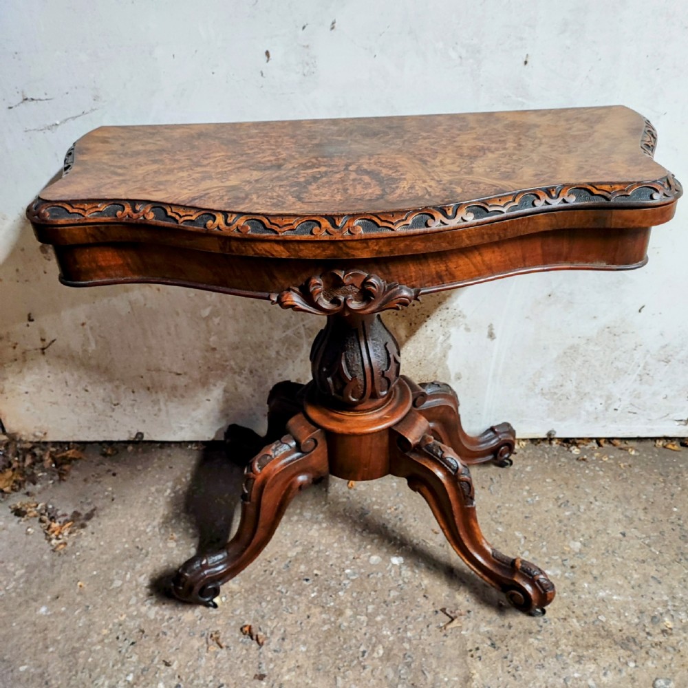 Victorian burr walnut fold-over card table featuring a serpentine top with carved apron details, supported by a central carved pedestal on four scroll-foot legs.