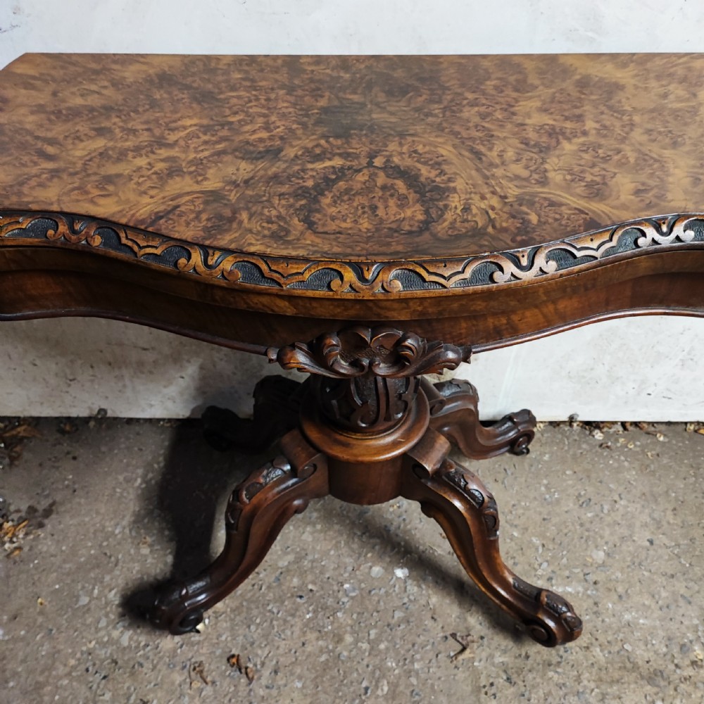 Victorian burr walnut fold-over card table featuring a serpentine top with carved apron details, supported by a central carved pedestal on four scroll-foot legs. View VI