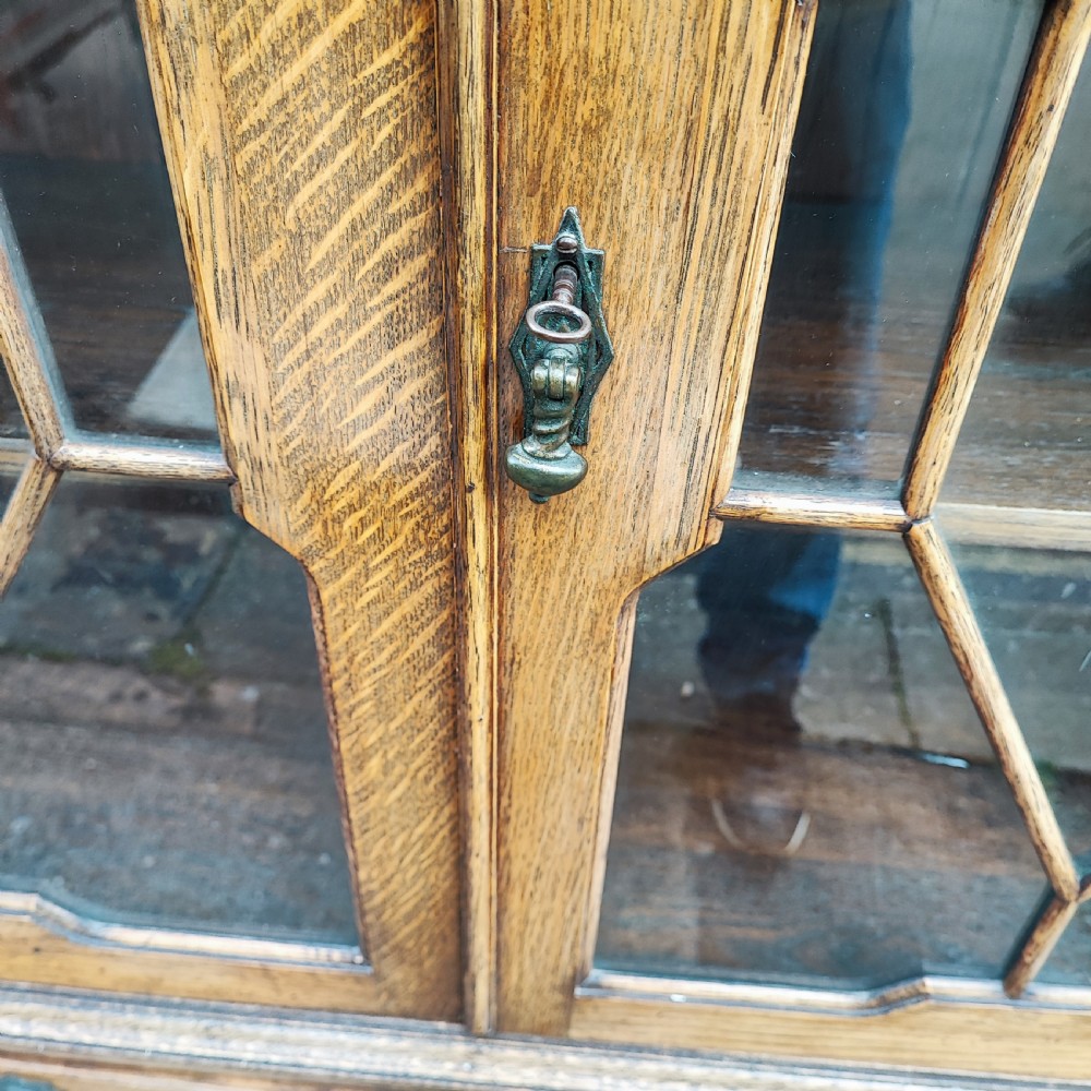 A warm brown oak Jacobean revival bookcase featuring double glazed doors with geometric diamond astragals, carved top frieze, and barley twist legs connected by a stretcher base. View III