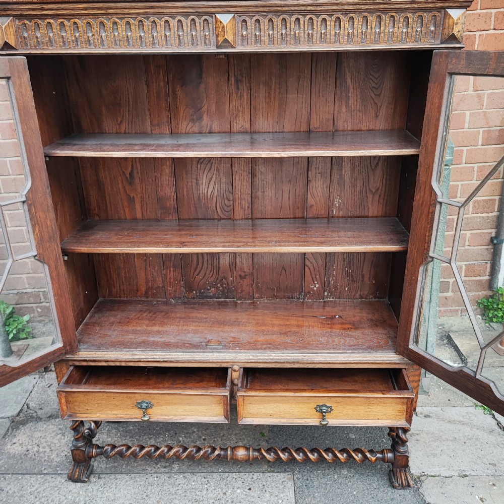 A warm brown oak Jacobean revival bookcase featuring double glazed doors with geometric diamond astragals, carved top frieze, and barley twist legs connected by a stretcher base. View VI