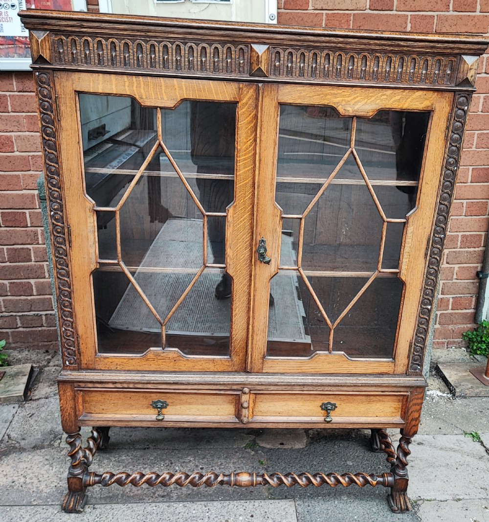 A warm brown oak Jacobean revival bookcase featuring double glazed doors with geometric diamond astragals, carved top frieze, and barley twist legs connected by a stretcher base. View VII
