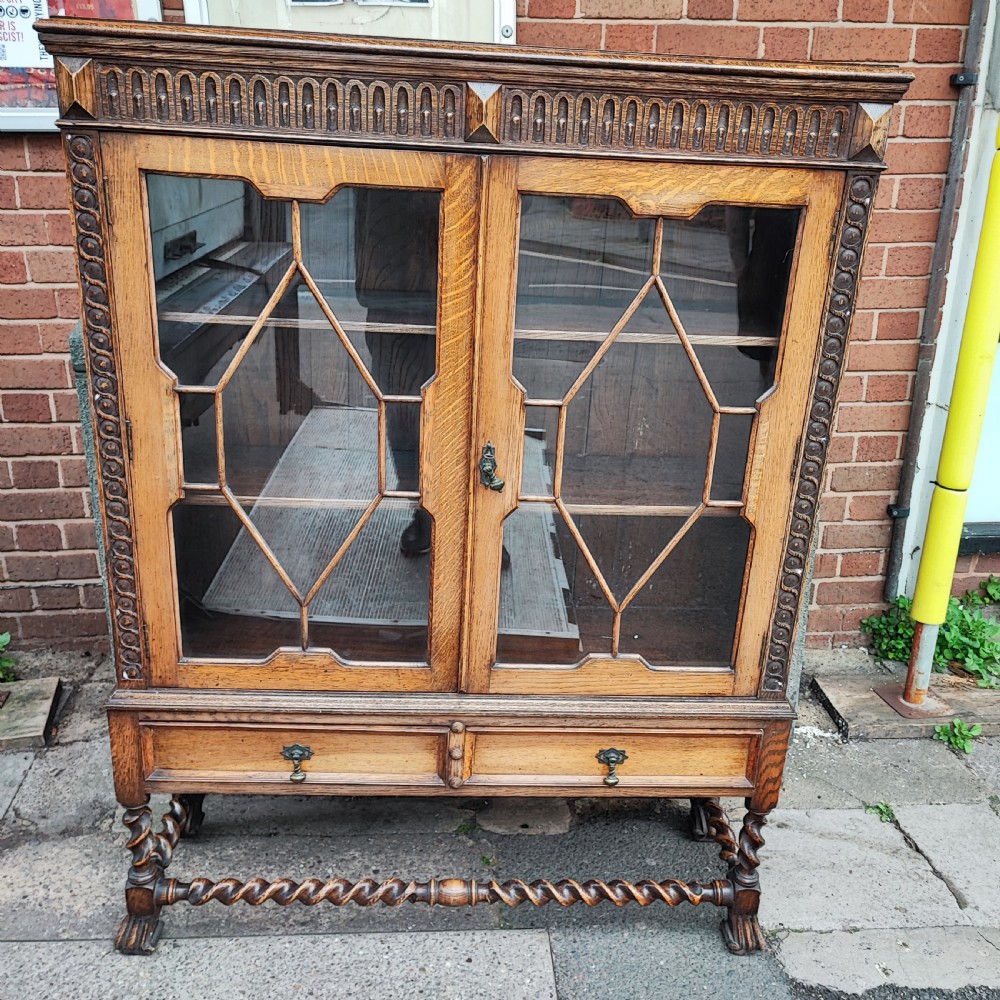 A warm brown oak Jacobean revival bookcase featuring double glazed doors with geometric diamond astragals, carved top frieze, and barley twist legs connected by a stretcher base. View VIII