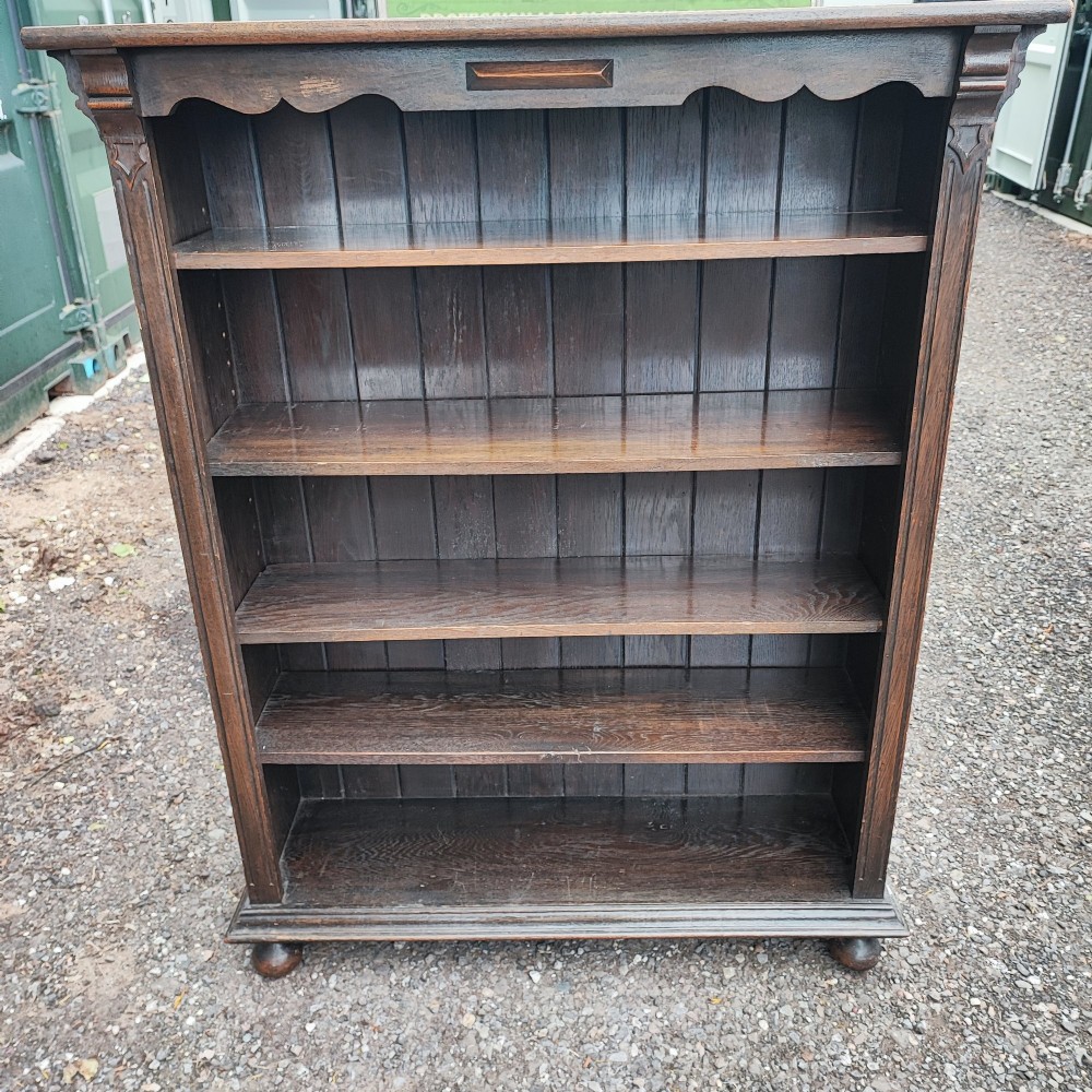 A dark oak open bookcase featuring five horizontal shelves, a scalloped upper frieze, vertical panelled backboards, and rounded bun feet.