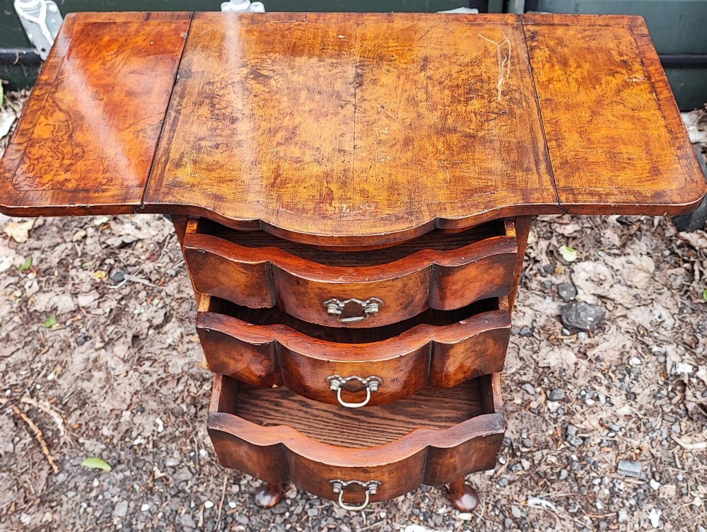 A warm brown walnut bedside chest featuring a serpentine front with three drawers, drop-leaf side flaps, brass handles, and slender cabriole legs. View II