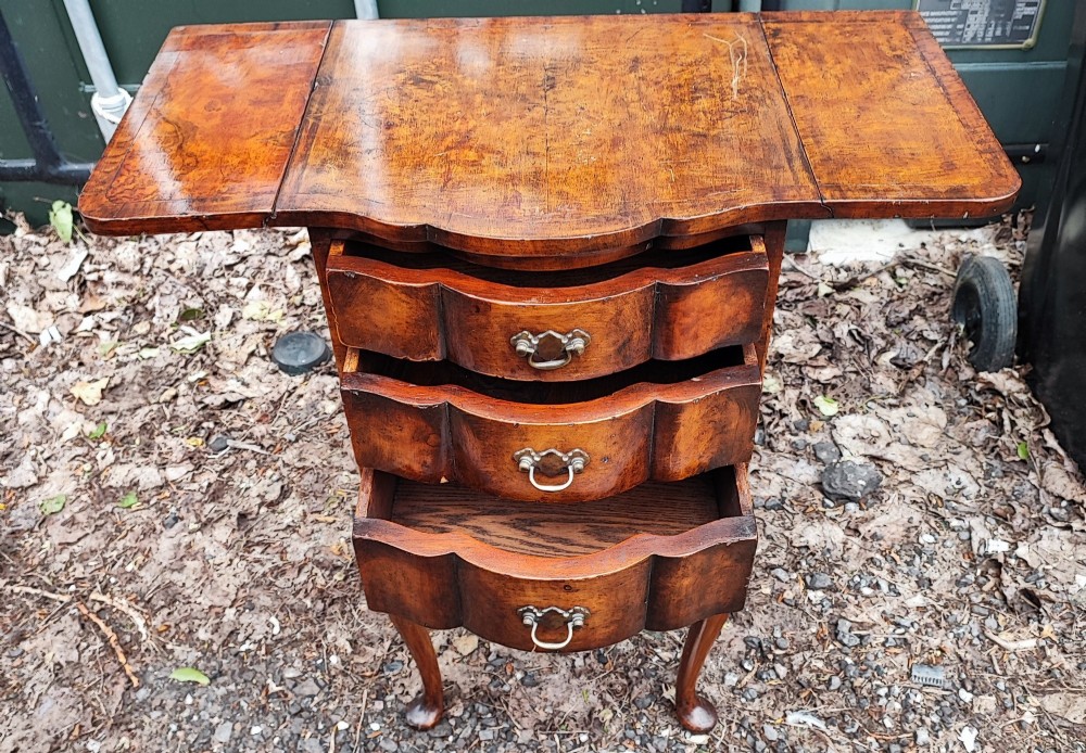 A warm brown walnut bedside chest featuring a serpentine front with three drawers, drop-leaf side flaps, brass handles, and slender cabriole legs. View IV