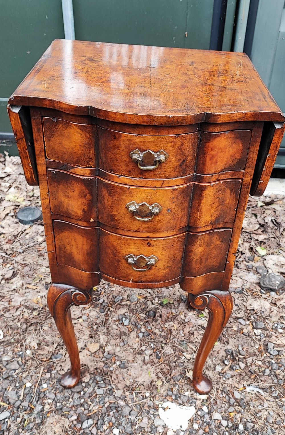 A warm brown walnut bedside chest featuring a serpentine front with three drawers, drop-leaf side flaps, brass handles, and slender cabriole legs. View V