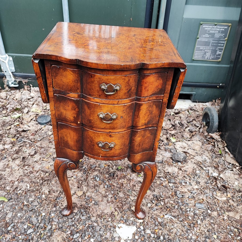 A warm brown walnut bedside chest featuring a serpentine front with three drawers, drop-leaf side flaps, brass handles, and slender cabriole legs. View VI