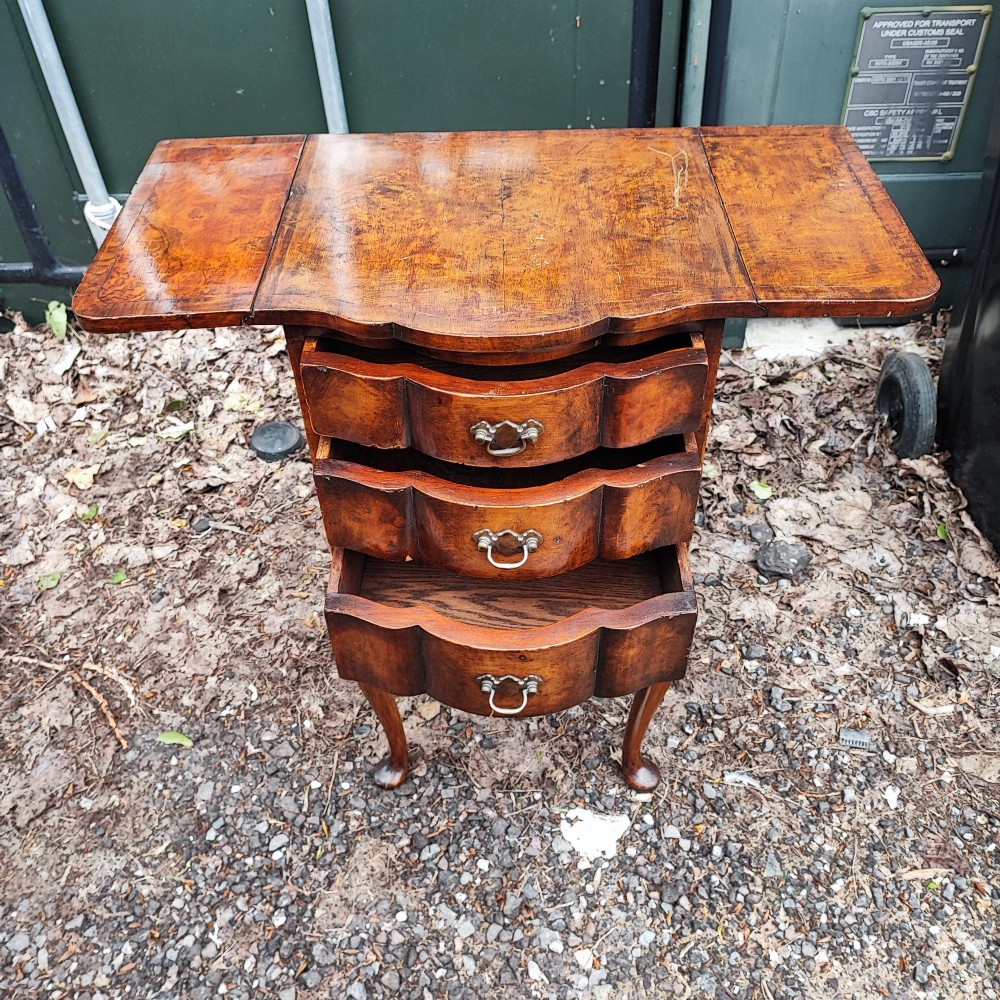 A warm brown walnut bedside chest featuring a serpentine front with three drawers, drop-leaf side flaps, brass handles, and slender cabriole legs. View VII
