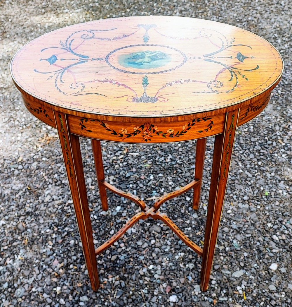 An oval painted satinwood occasional table with a central cherub motif, floral swags, tapered legs, and an X-shaped stretcher.