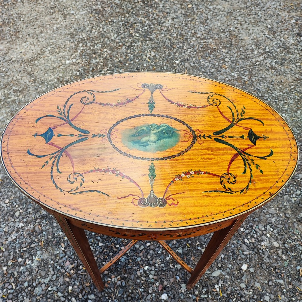 An oval painted satinwood occasional table with a central cherub motif, floral swags, tapered legs, and an X-shaped stretcher. View II
