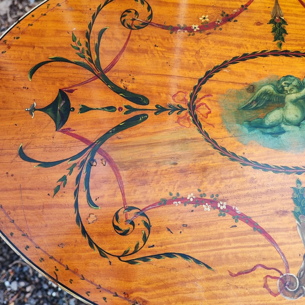 An oval painted satinwood occasional table with a central cherub motif, floral swags, tapered legs, and an X-shaped stretcher. View III