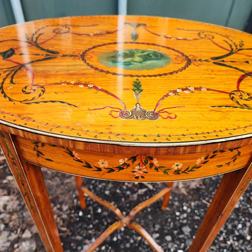 An oval painted satinwood occasional table with a central cherub motif, floral swags, tapered legs, and an X-shaped stretcher. View V