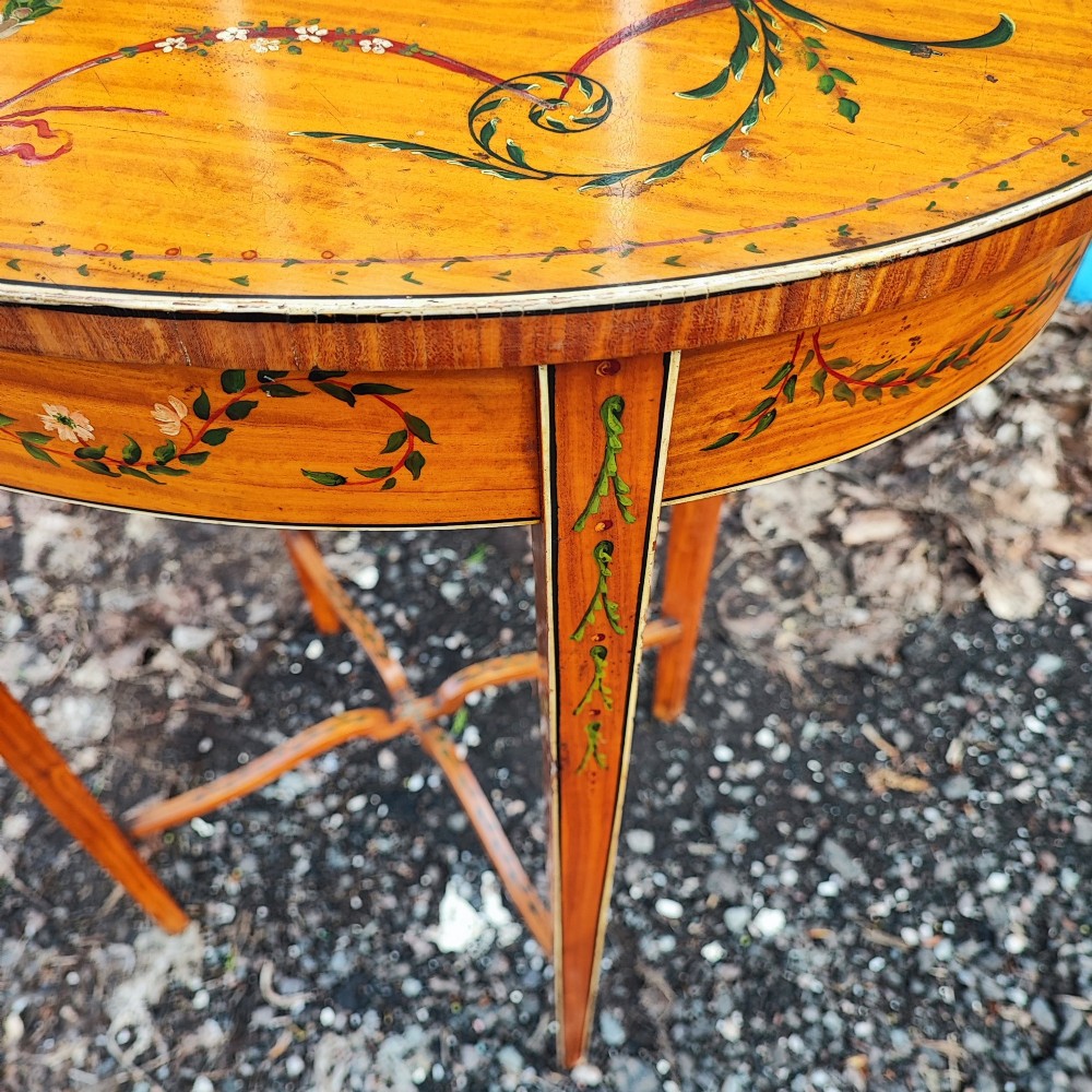 An oval painted satinwood occasional table with a central cherub motif, floral swags, tapered legs, and an X-shaped stretcher. View VI