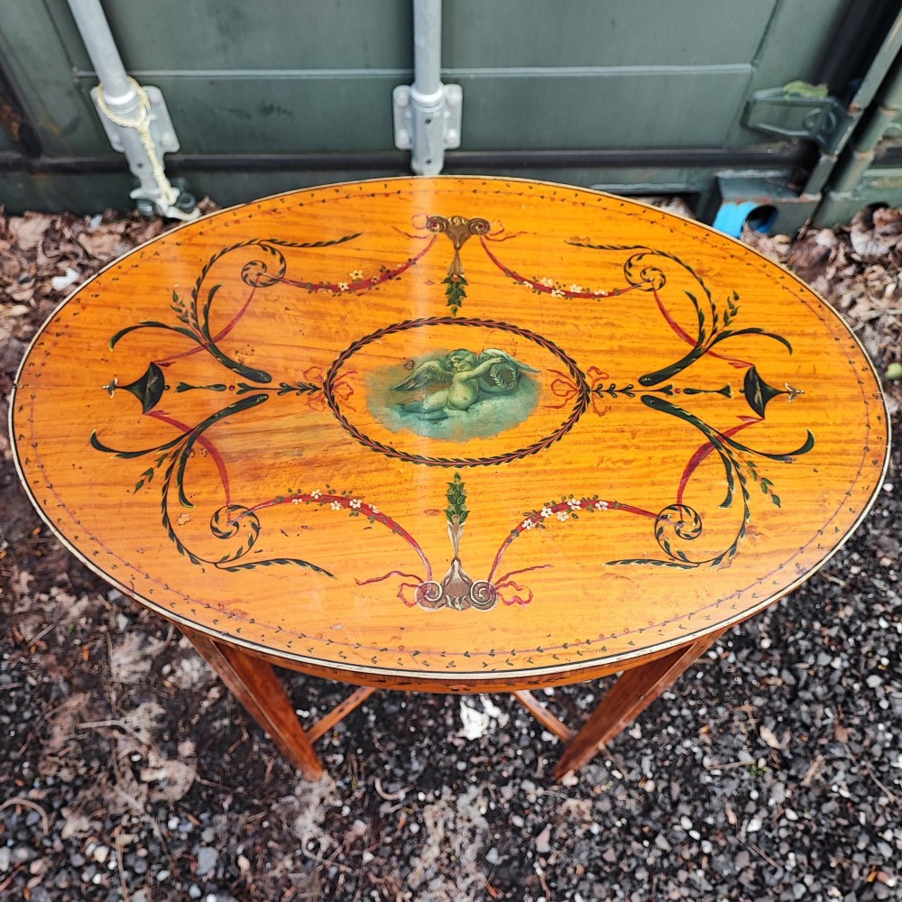 An oval painted satinwood occasional table with a central cherub motif, floral swags, tapered legs, and an X-shaped stretcher. View VIII