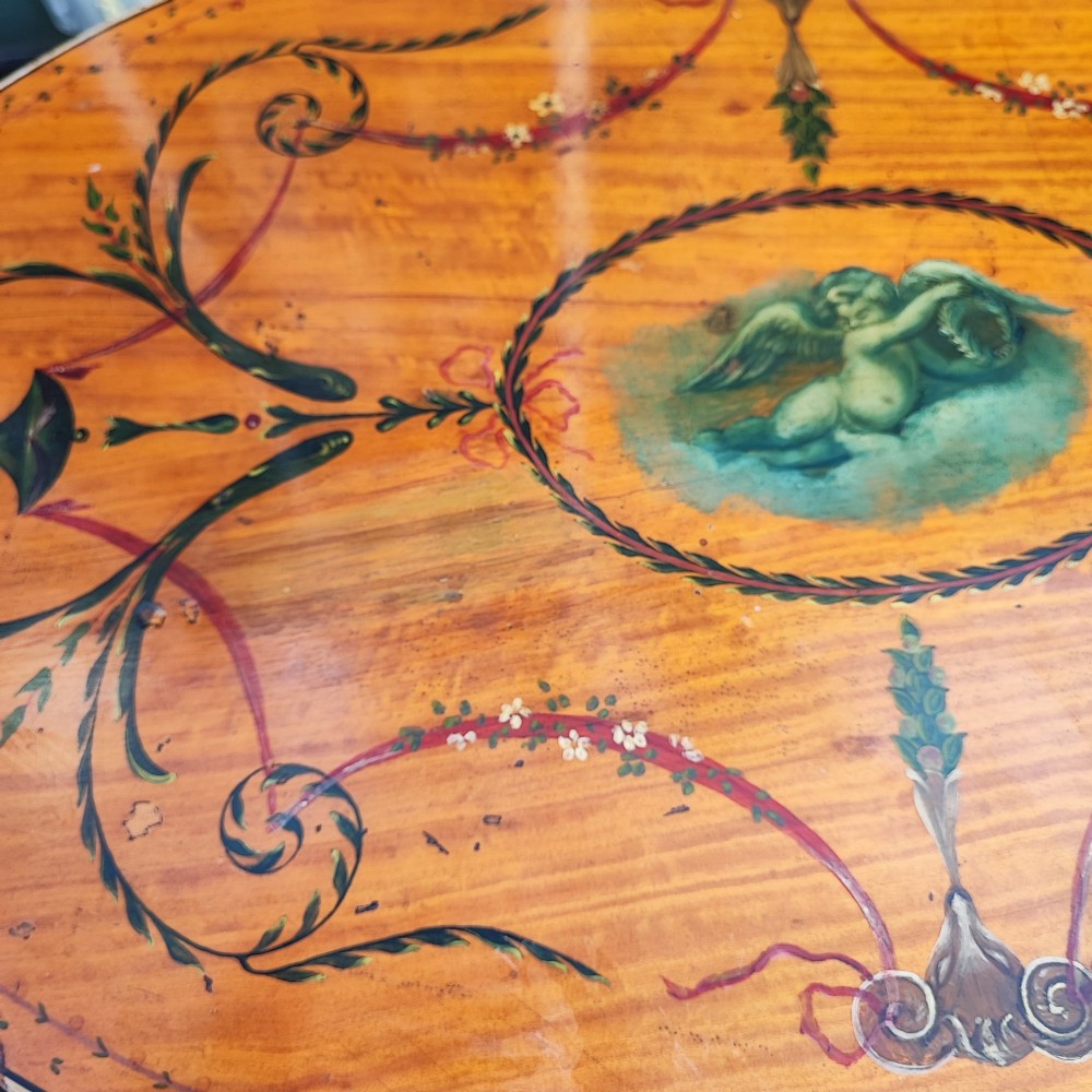 An oval painted satinwood occasional table with a central cherub motif, floral swags, tapered legs, and an X-shaped stretcher. View IX