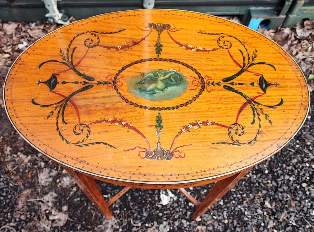 An oval painted satinwood occasional table with a central cherub motif, floral swags, tapered legs, and an X-shaped stretcher. View X
