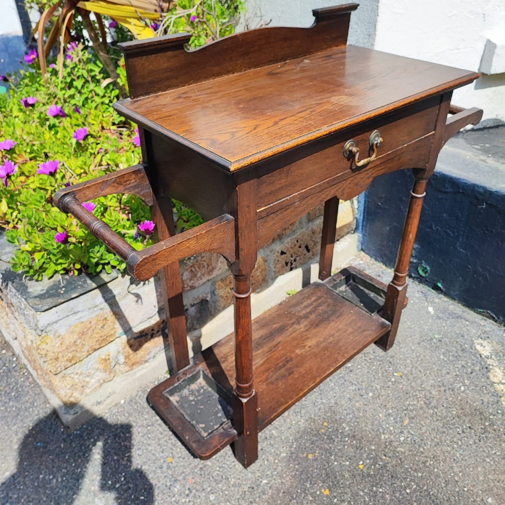 An Arts and Crafts oak hall stick stand featuring a flat top table, single drawer, side umbrella rails, turned legs, and metal drip trays on the lower shelf. View II