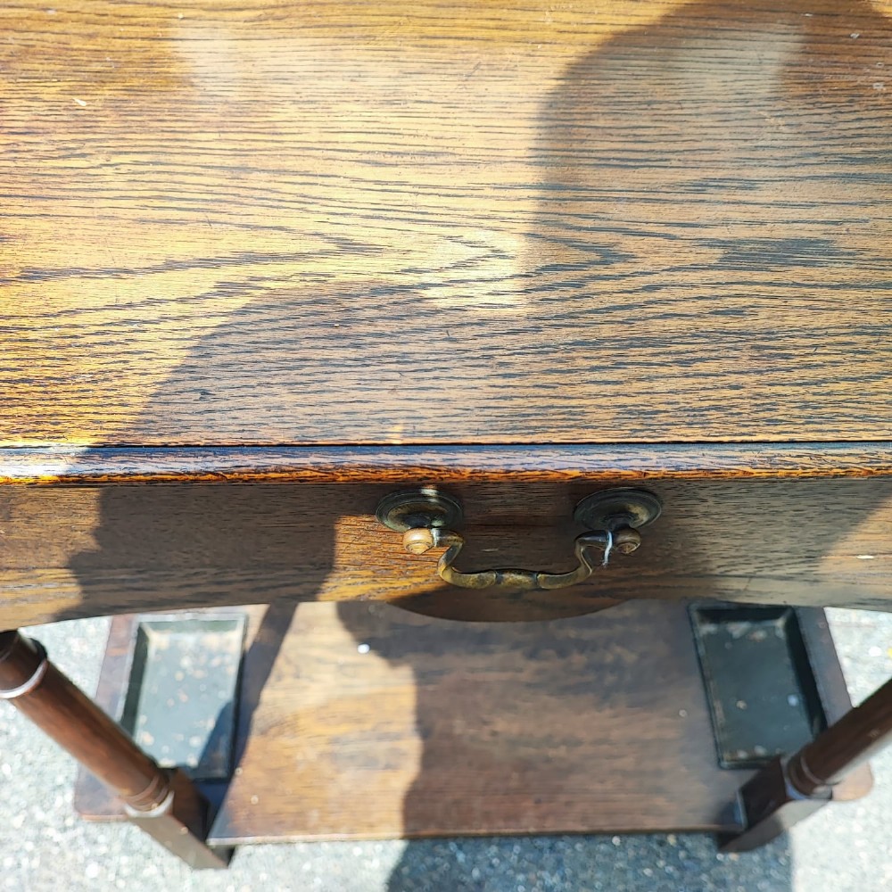 An Arts and Crafts oak hall stick stand featuring a flat top table, single drawer, side umbrella rails, turned legs, and metal drip trays on the lower shelf. View III
