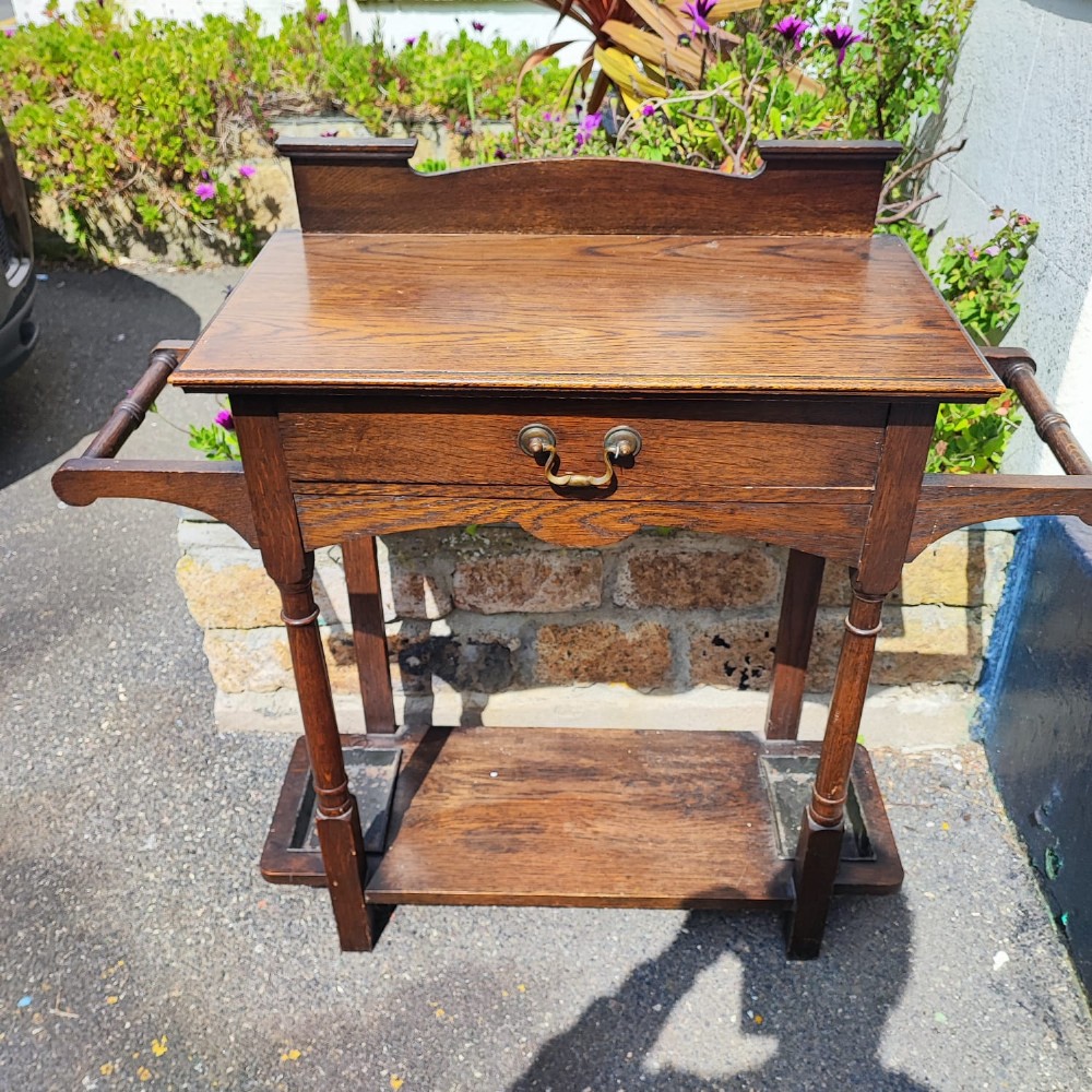 An Arts and Crafts oak hall stick stand featuring a flat top table, single drawer, side umbrella rails, turned legs, and metal drip trays on the lower shelf. View IV