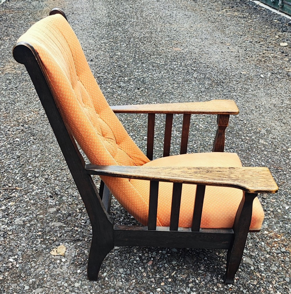An antique Arts and Crafts oak reclining armchair with broad flat armrests, slatted sides, and orange buttoned upholstery.
