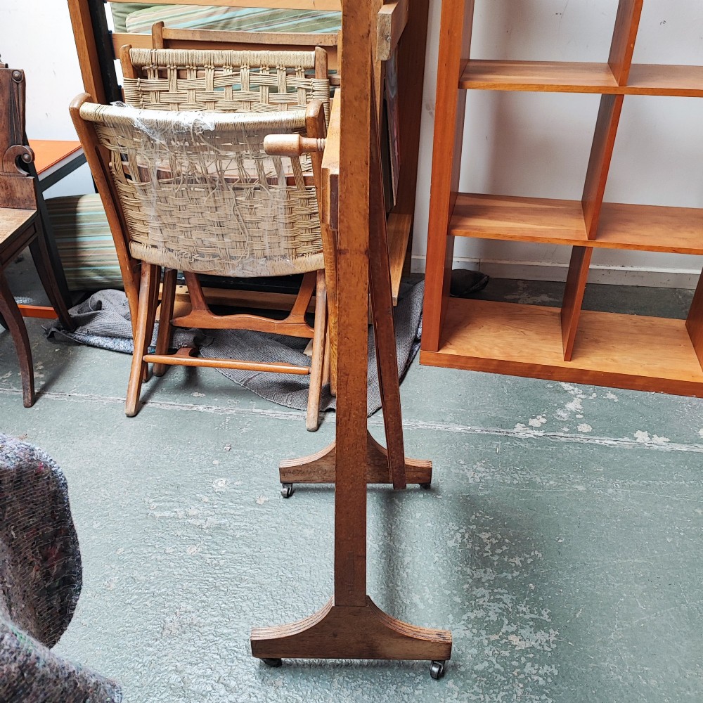 An Arts and Crafts oak cheval mirror featuring a tall bevelled glass plate supported between two square upright posts on splayed legs with castors. View II