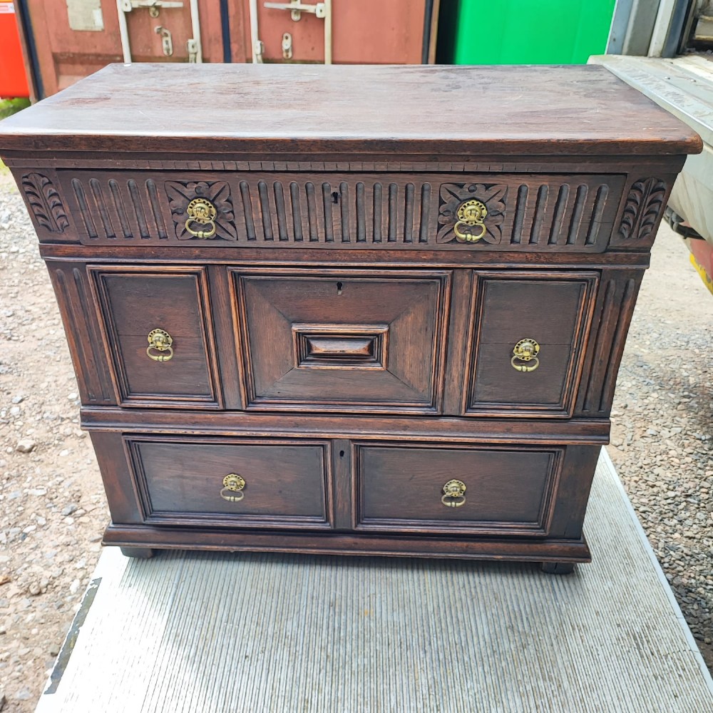 A dark oak Jacobean chest of drawers featuring geometric panel moulding, a carved top drawer, and brass drop handles. View VI