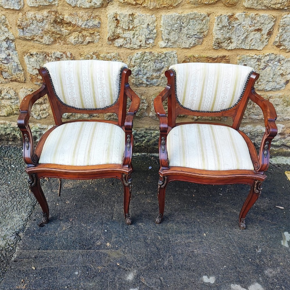 A matching pair of French antique armchairs crafted from polished brown wood with curved arms, cabriole legs, and cream striped fabric upholstery with studded borders.