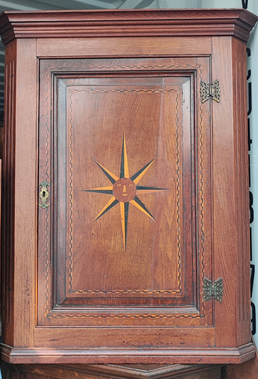 An antique brown oak hanging corner cupboard featuring a central sunburst inlaid door panel, chequered border stringing, and decorative brass hinges. View IV