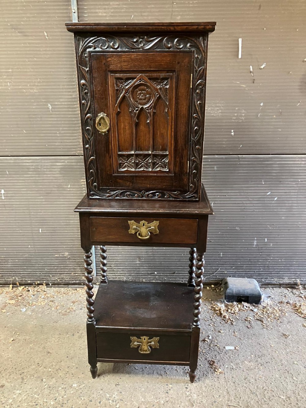 An antique dark carved oak hall cabinet featuring an upper cupboard door with Gothic arched relief carving, two drawers with brass drop handles, and turned barley twist legs. View II