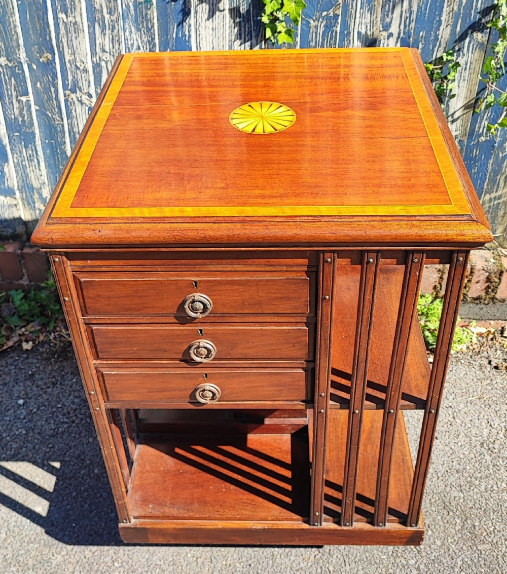 An Edwardian inlaid mahogany revolving bookcase featuring a square top with a central starburst patera inlay, three small drawers with brass ring handles, and vertical timber side slats.
