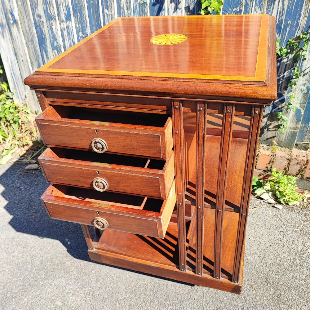 An Edwardian inlaid mahogany revolving bookcase featuring a square top with a central starburst patera inlay, three small drawers with brass ring handles, and vertical timber side slats. View IV