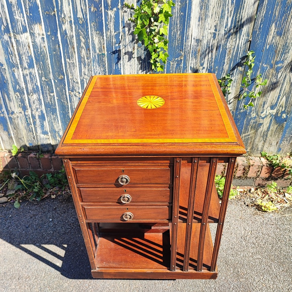 An Edwardian inlaid mahogany revolving bookcase featuring a square top with a central starburst patera inlay, three small drawers with brass ring handles, and vertical timber side slats. View IX
