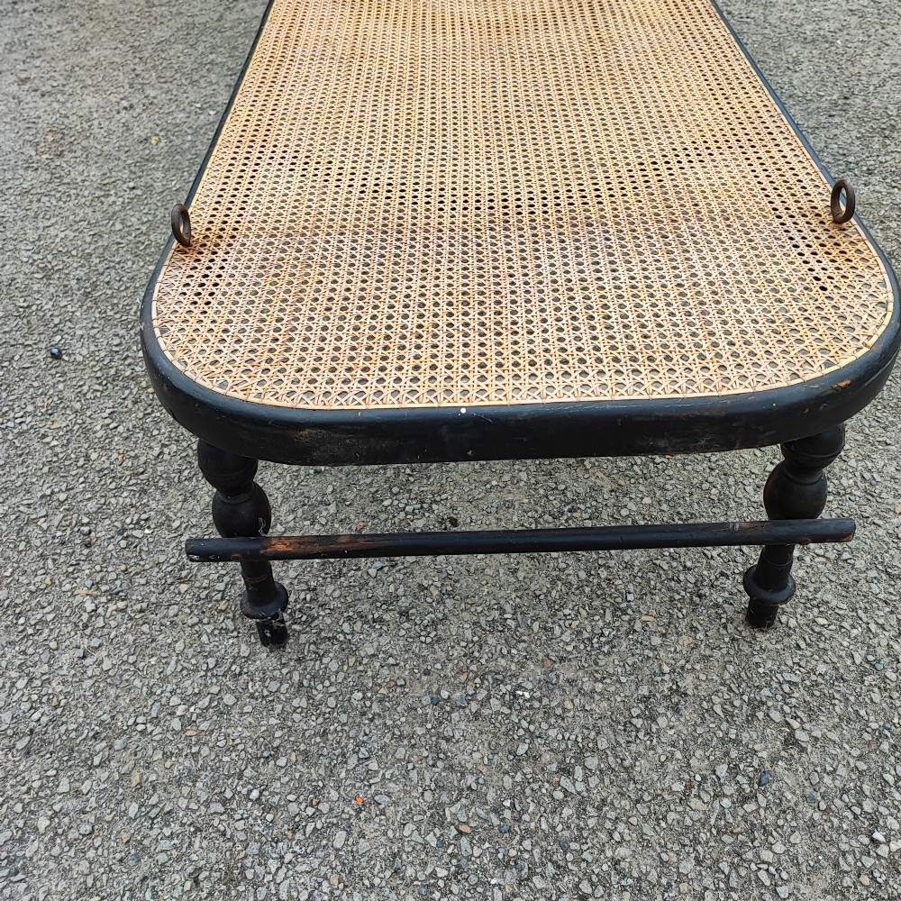 An ebonised wooden daybed with a long hand-woven cane seat, turned legs, and an integrated low headrest. View VI