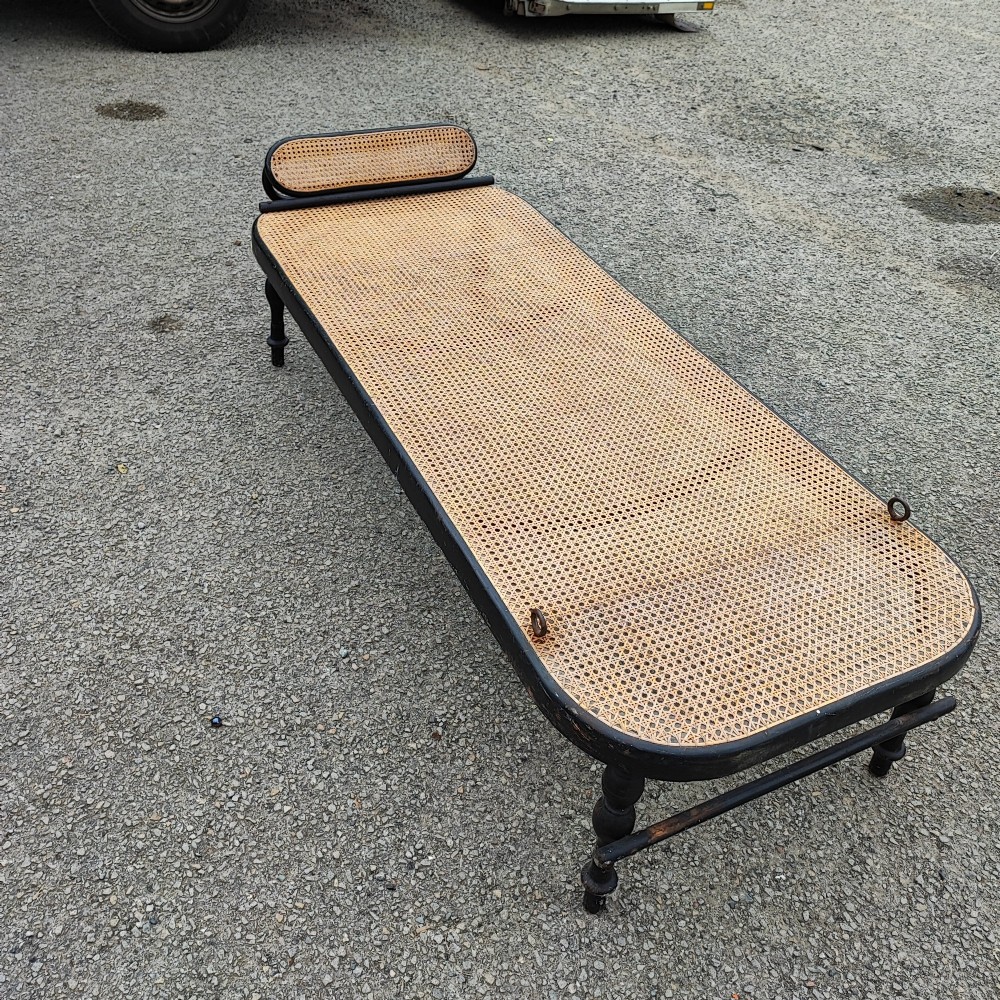 An ebonised wooden daybed with a long hand-woven cane seat, turned legs, and an integrated low headrest. View VII