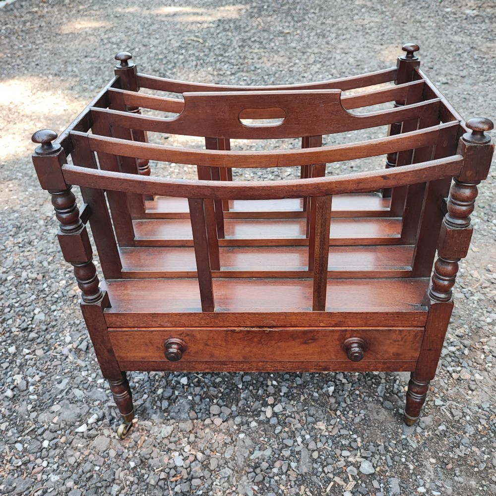 An antique Regency mahogany Canterbury featuring slatted vertical dividers, a central carrying handle, a single base drawer, and turned legs with brass castors.