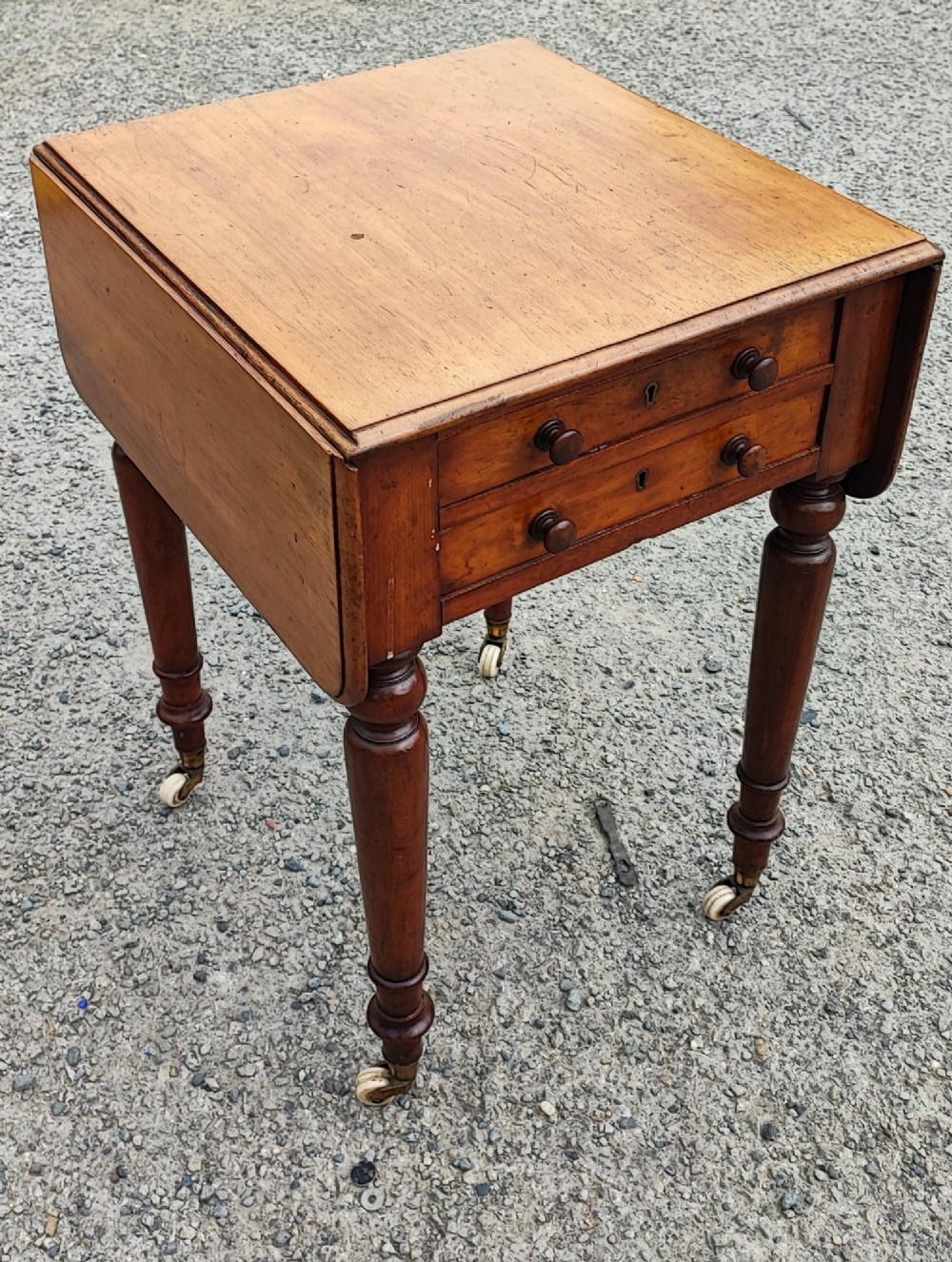 A warm brown mahogany Pembroke table featuring two drop-down leaves, two front drawers with round knobs, and turned legs fitted with castors.