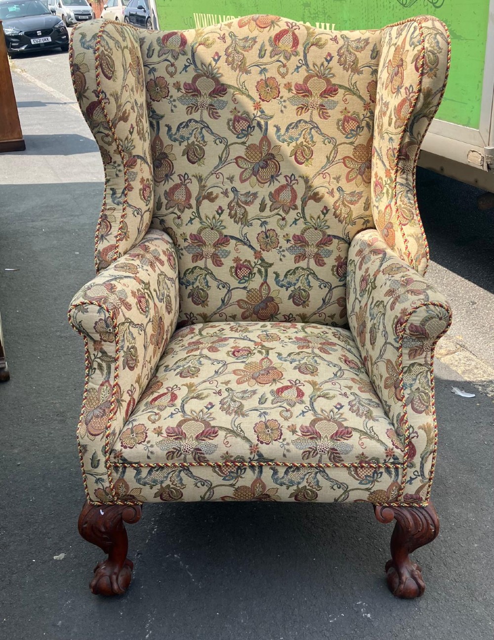A high-back wing chair featuring floral tapestry upholstery, rolled arms, braided edge trim, and carved mahogany cabriole front legs with ball and claw feet.