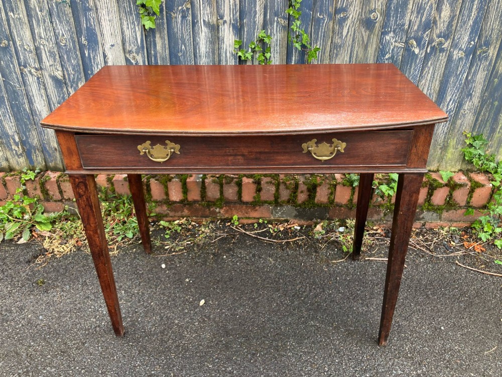 A Georgian bow-fronted mahogany side table featuring a single drawer with two brass handles and supported on four square tapered legs.